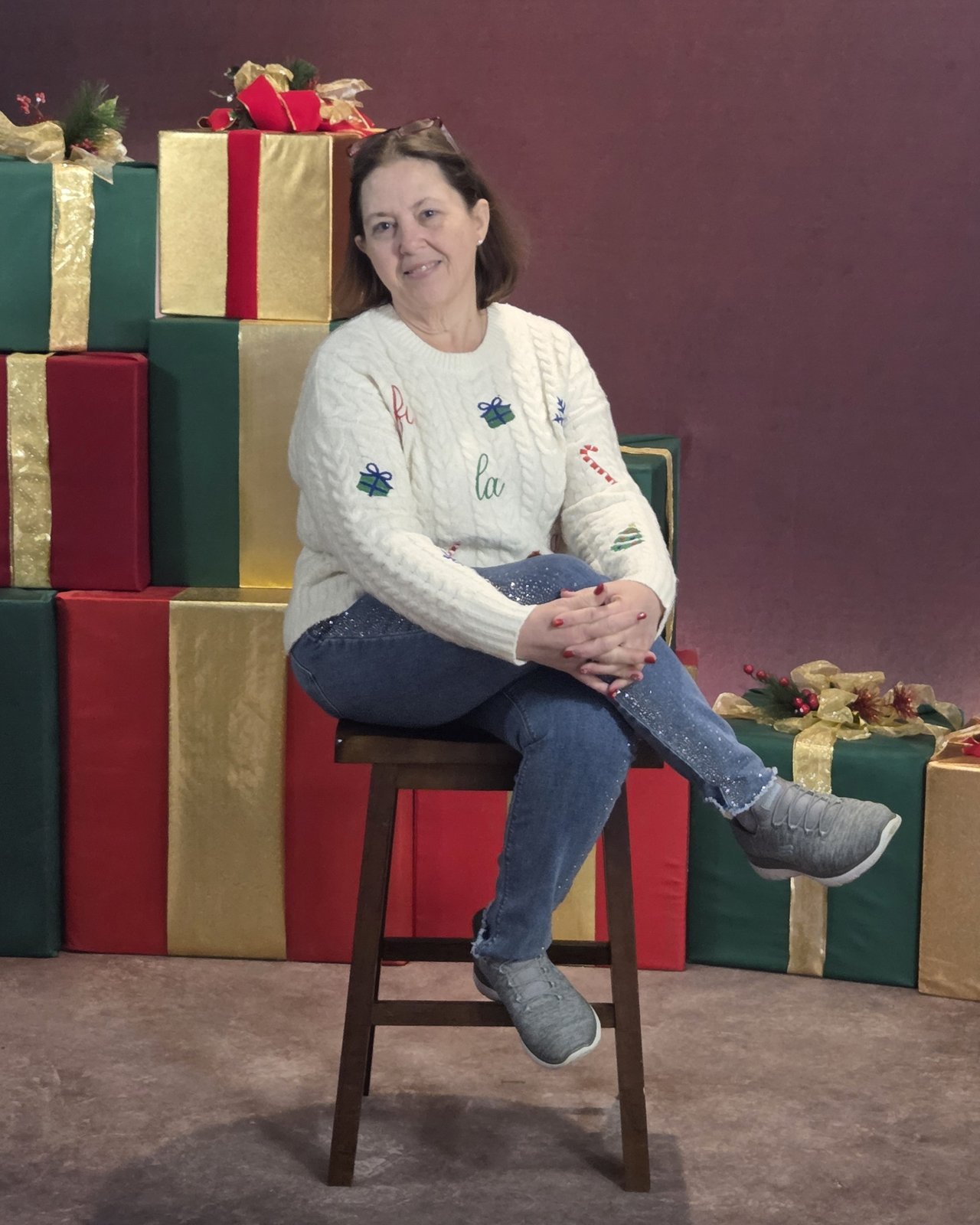 Wanda seated on a stool in a cream Christmas sweater, with stacks of wrapped gifts beside her